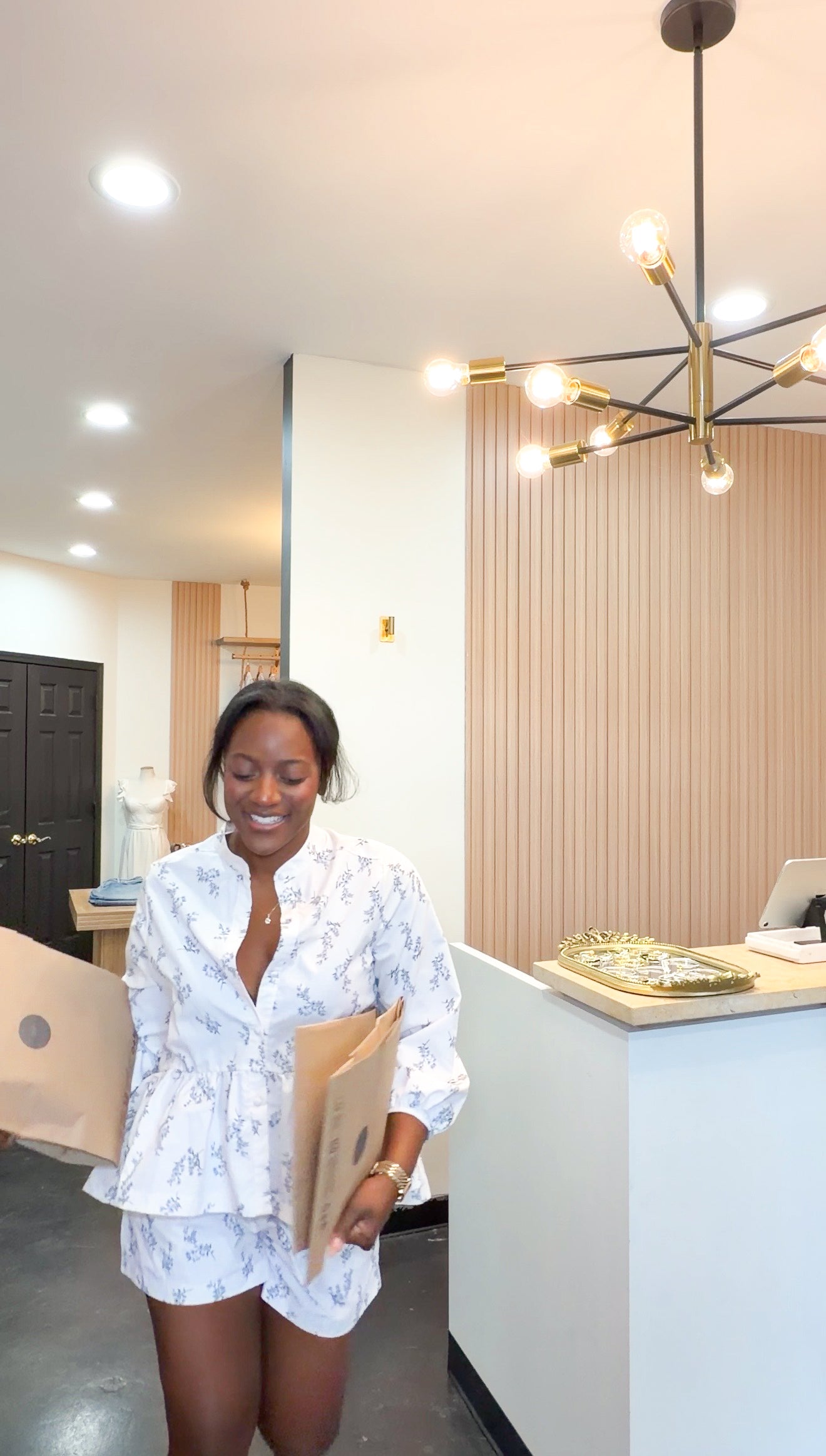 A woman wearing a white short-sleeve button-up shirt and matching shorts with a delicate blue floral pattern, holding cardboard boxes in a modern boutique setting.